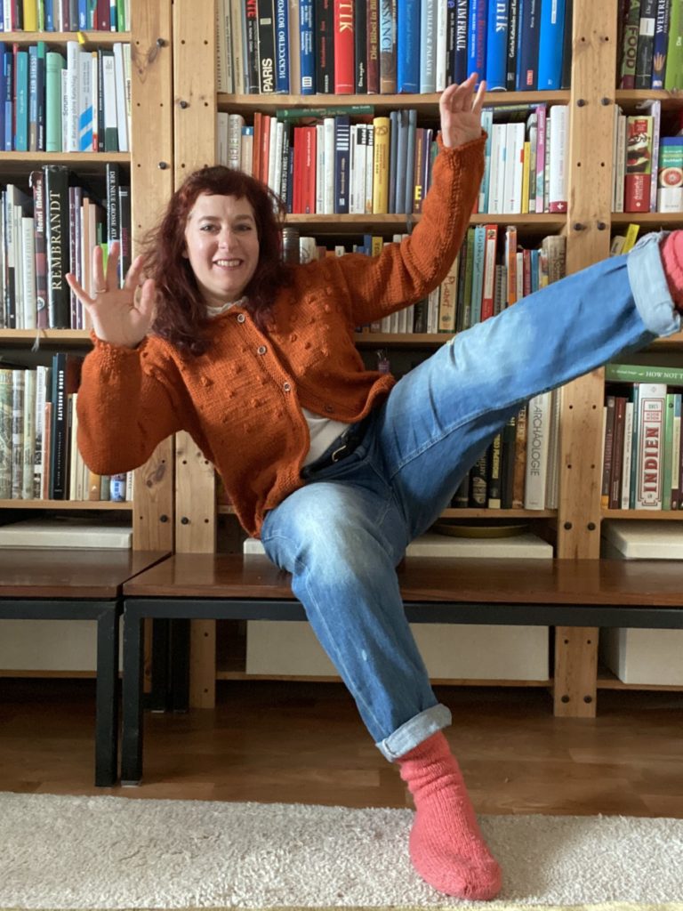 woman wearing a orange colored hand knitted Rönn cardigan in forn of a huge book shelf